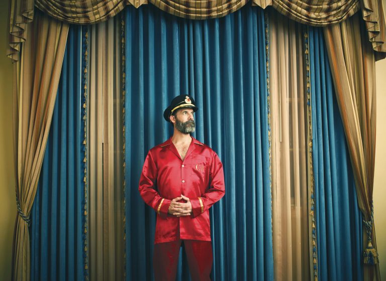 Portrait photograph of funny Captain Obvious with his hands clasped while staring into the distance. Captain Obvious is wearing a red silk suit standing in front of elegant blue and gold curtains. 