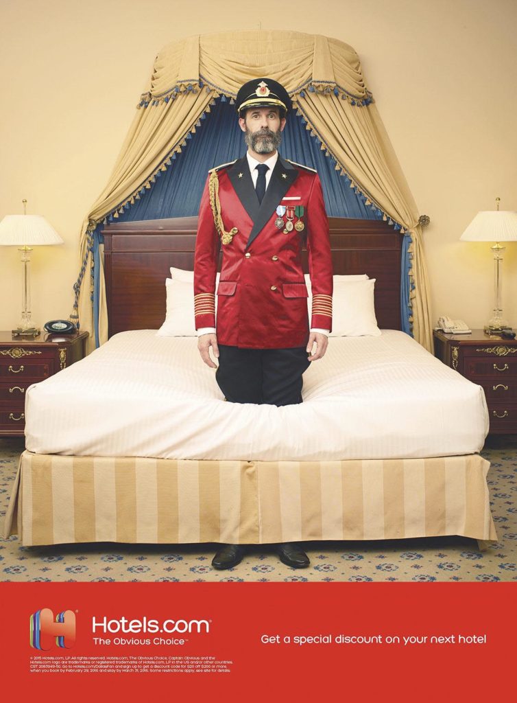Funny advertisement of Captain Obvious standing through a bed in a hotel room while wearing a red captain suit and hat. Funny still photography campaign for Hotels.com photographed by The Wade Brothers.