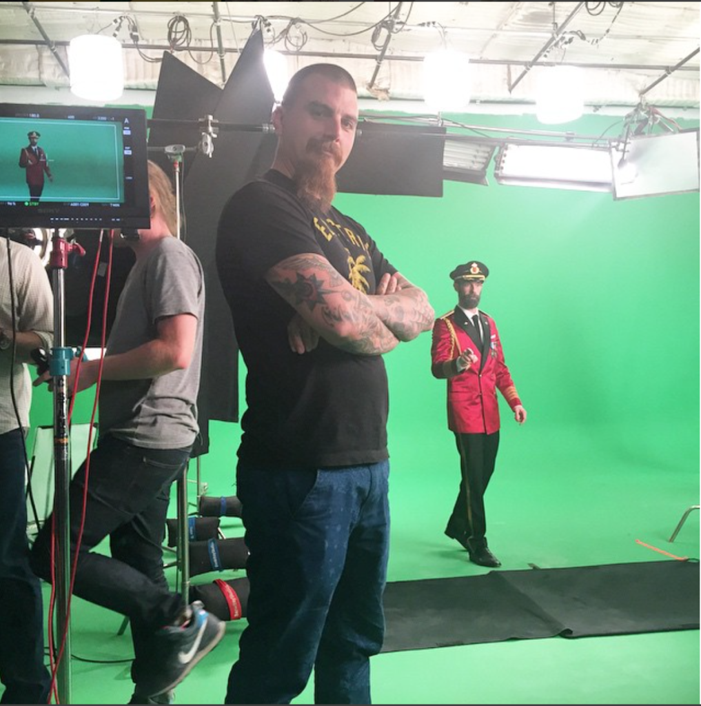Funny behind-the-scenes photograph of Lindsey Wade proudly standing in front of camera gear while on set of Captain Obvious still photography ad campaign. Captain Obvious stands in front of a green screen in the background. 