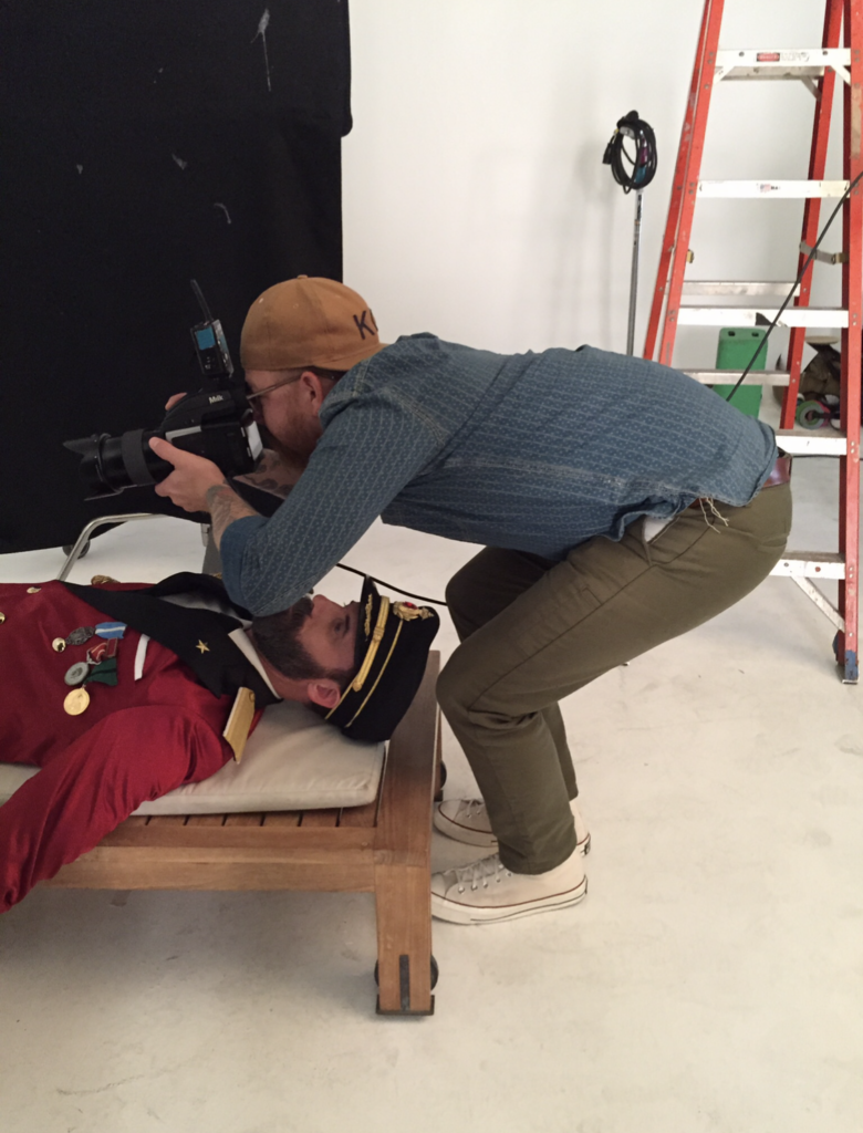 Behind the scenes photograph of Lyndon Wade creating still photography for The Obvious Choice campaign for Hotels.com with viral brand mascot Captain Obvious. Lyndon Wade crouching over Captain Obvious capturing still photography of Captain Obvious' legs and feet. 